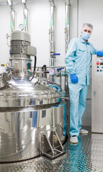 A man in a blue Lab Coat stands beside a large cosmetic manufacturing machine